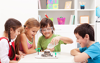 2 boys and 2 girls gathered around a table while one boy is pouring water into a structure to make a volcano